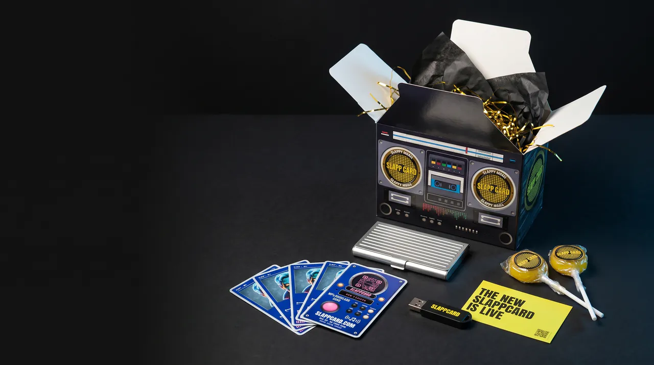 A boombox-printed box standing open on a dark table, with Slappcards, a metal card holder, a USB stick, two suckers and a yellow card laid out in front of it